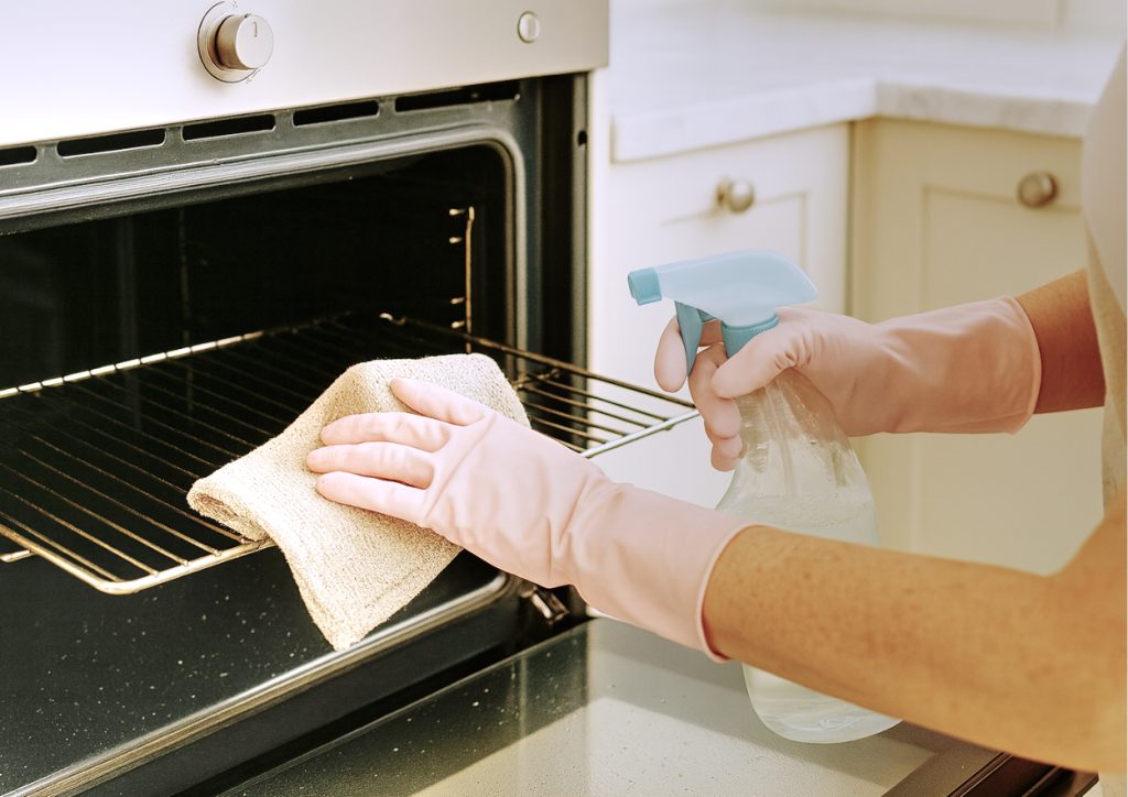 An image of an unseen person wearing pale pink rubber gloves, cleaning an oven using a clean microfibre cloth and a solution of eco-friendly bicarbonate of soda, vinegar and lemon cleaning solution.
