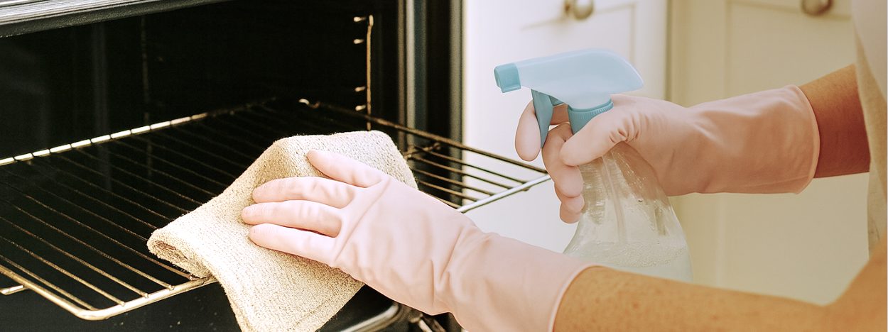 An image of an unseen person wearing pale pink rubber gloves, cleaning an oven using a clean microfibre cloth and a solution of eco-friendly bicarbonate of soda, vinegar and lemon cleaning solution.